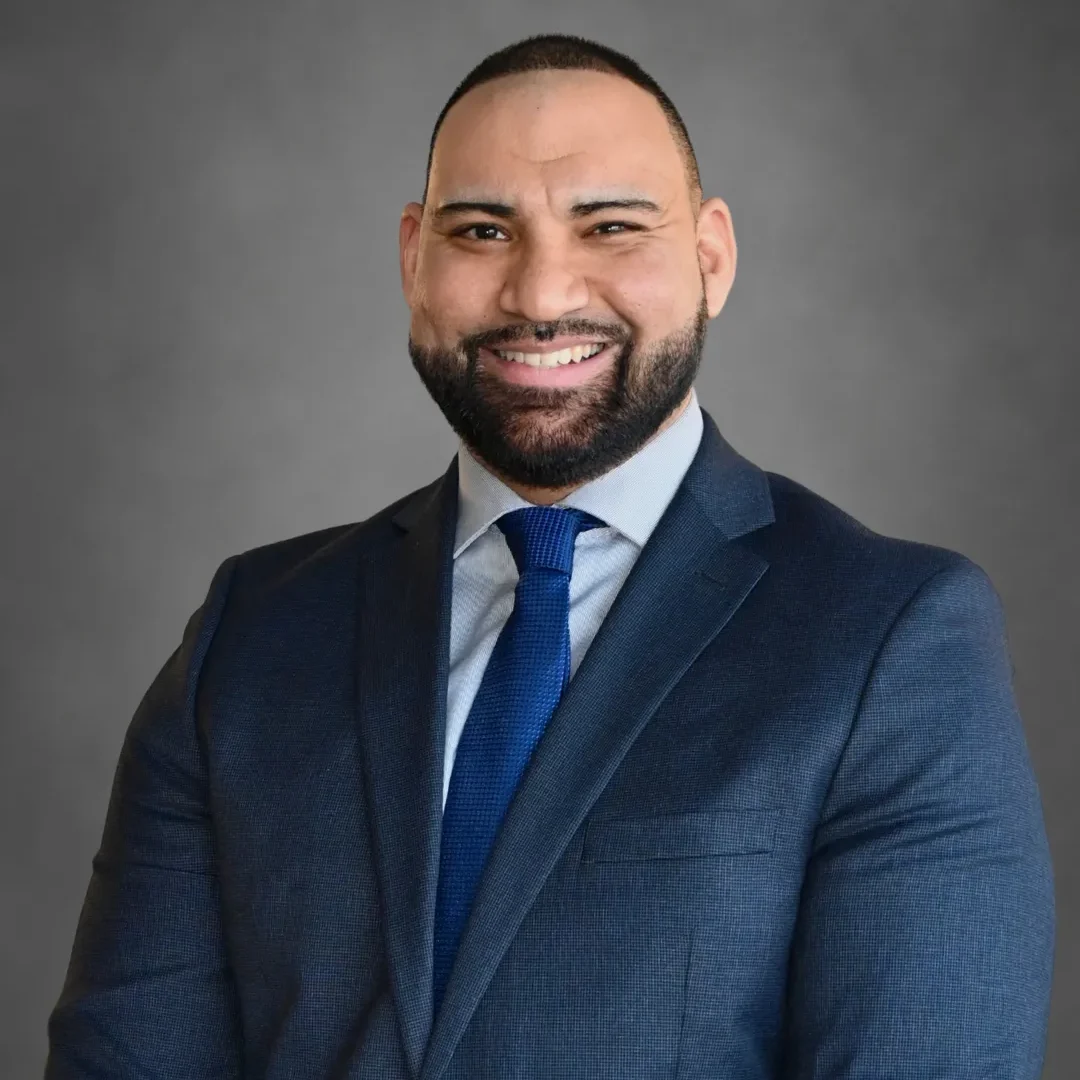Professional headshot of a smiling man with a beard, wearing a dark blue suit and tie against a gray background.