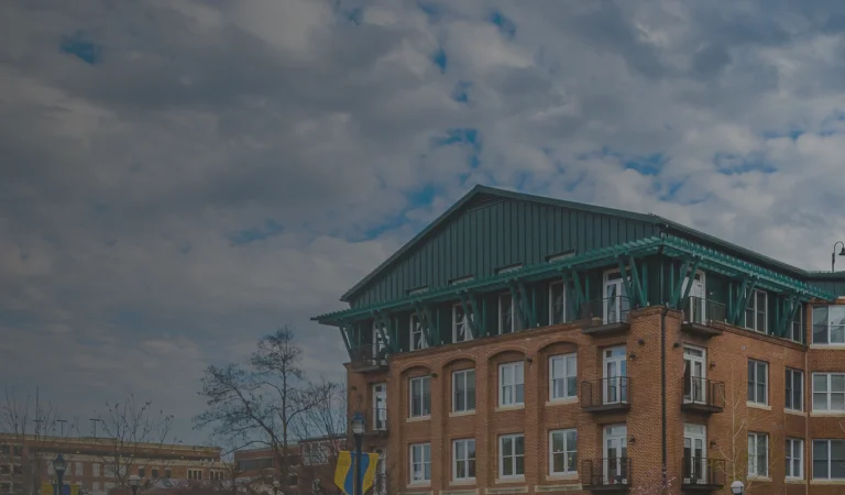 A row of red brick buildings with green roofs under a cloudy sky, a light post in the foreground, and yellow banners on poles.