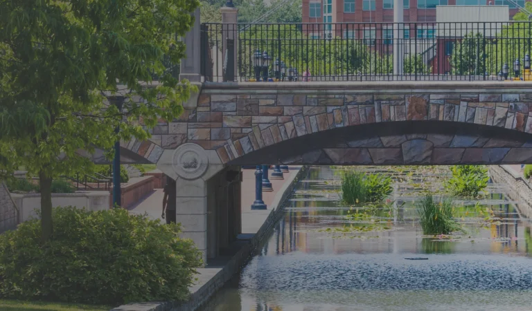 A panoramic view of a stone bridge over a canal surrounded by greenery and street lamps, with a person walking under the bridge.