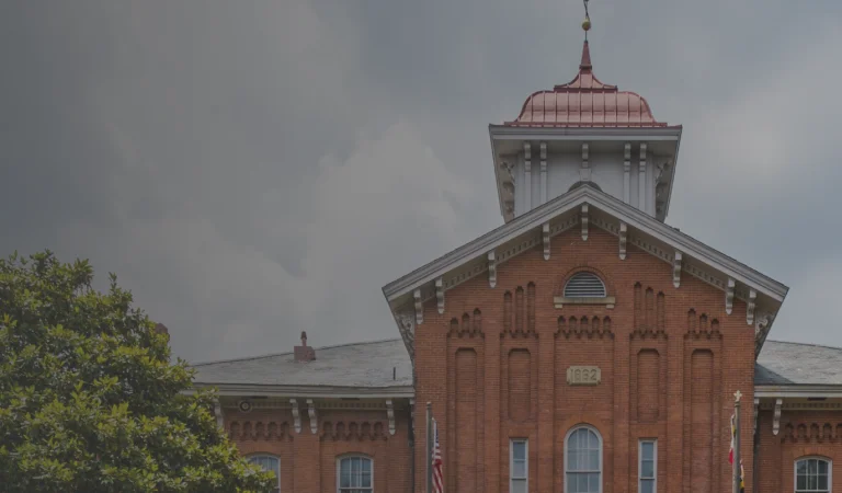 Old red brick building with a tall bell tower and white cupola against a cloudy sky, surrounded by green trees.