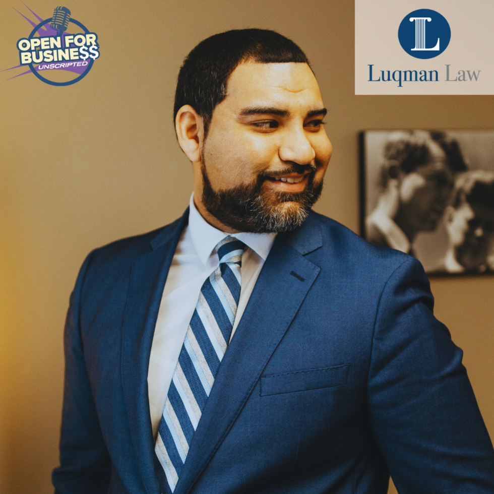 Smiling bearded man in a dark blue suit and blue striped tie, with logos for "Open For Business Unscripted" and "Luqman Law" in the corners.
