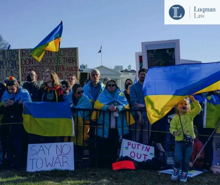 Group of people at an outdoor rally holding Ukrainian flags and protest signs, some with messages against Putin.