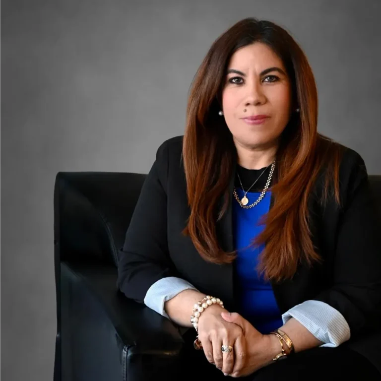 Professional woman with long brown hair, wearing a black blazer and blue top, seated on black chair.