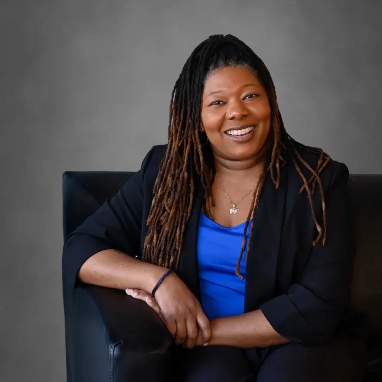Professional portrait of a smiling woman with long dreadlocks, wearing a blue top and black blazer.